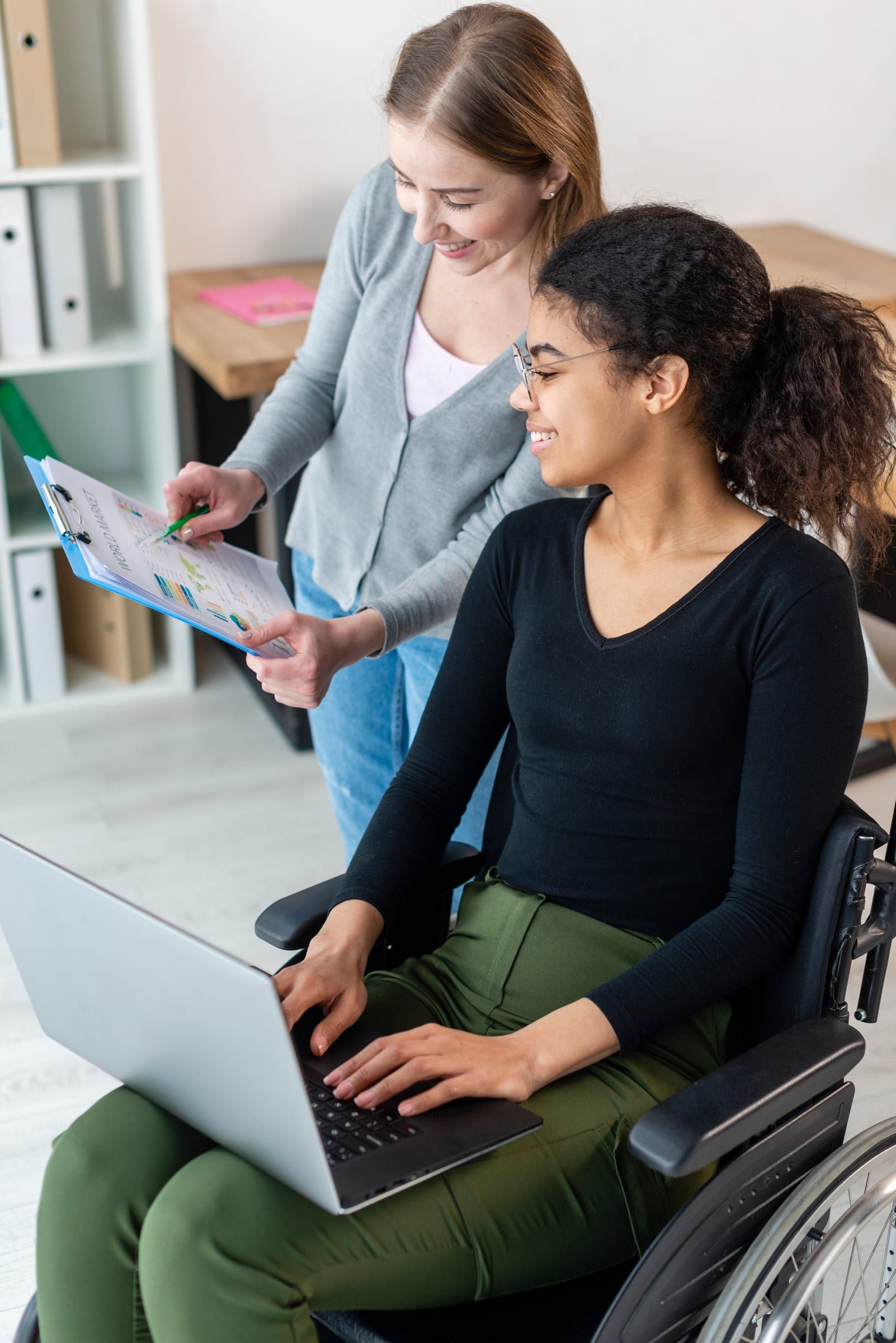A woman using a wheelchair works on a laptop while another woman points to notes on a clipboard