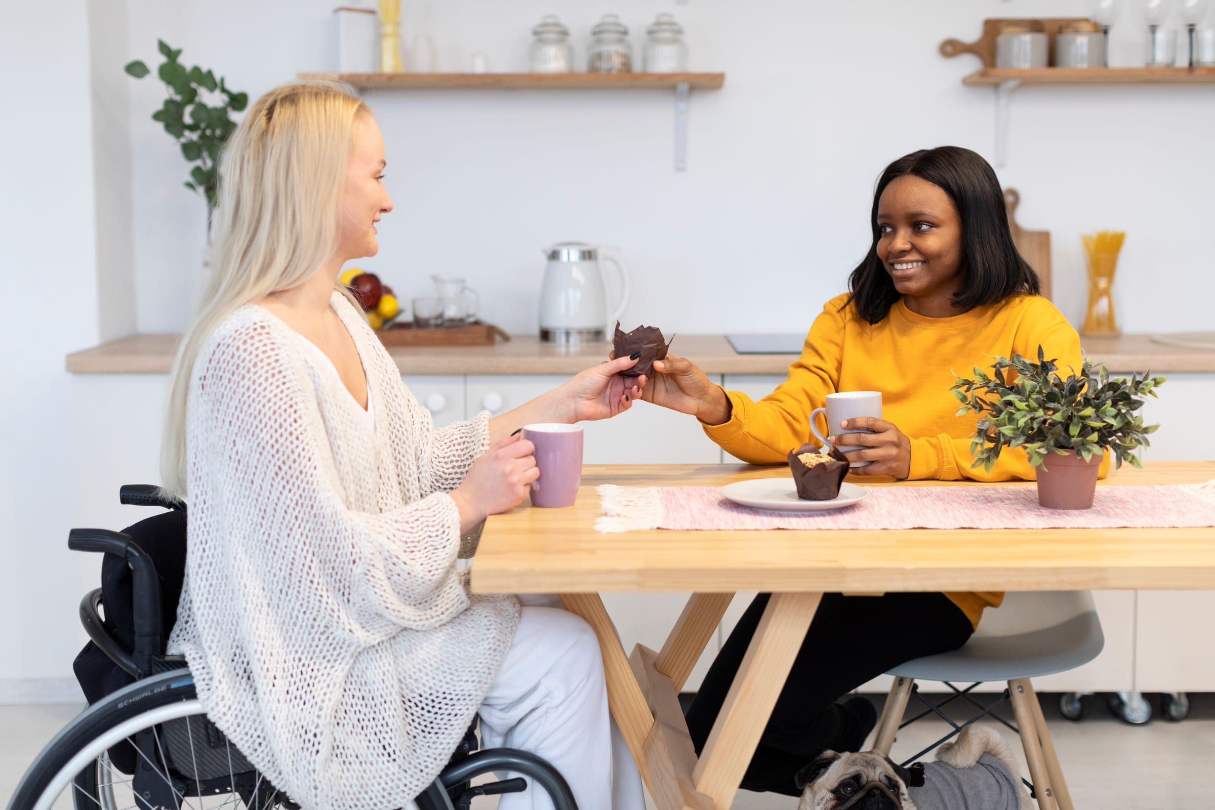 A woman using a wheelchair shares morning tea and muffins with a friend at a kitchen table